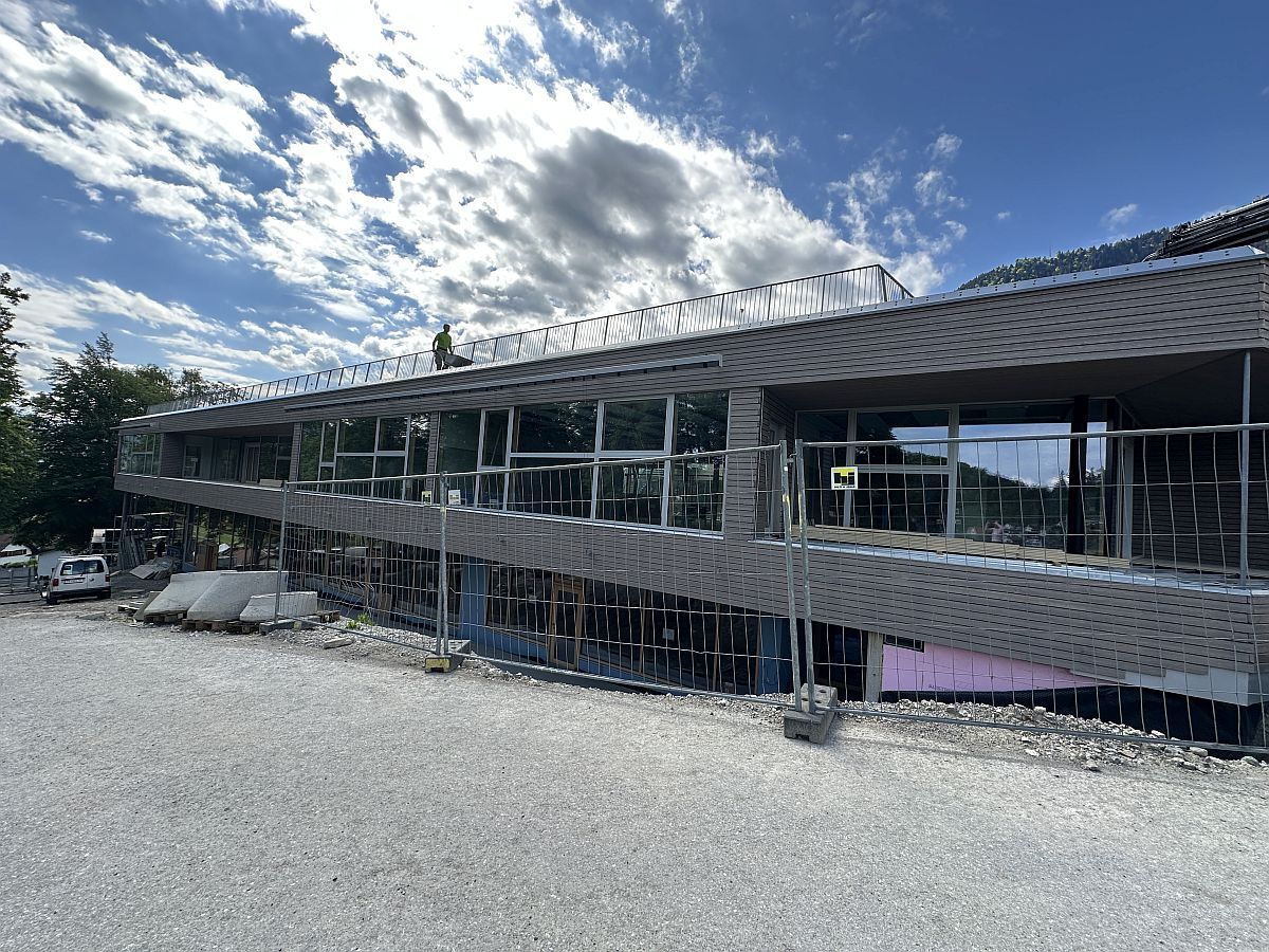 Ein modernes Gebäude im Bau mit Glaswänden und einer Person auf dem Dach, umgeben von Zäunen und Baumaterialien, mit einem bewölkten Himmel im Hintergrund.