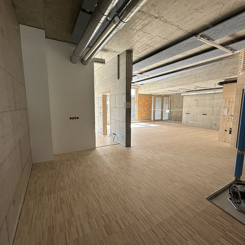 Ein leerer Raum mit Holzböden und sichtbaren Betonwänden. Rohre sind an der Decke montiert. Es gibt zwei Türöffnungen, die zu anderen Räumen führen.
