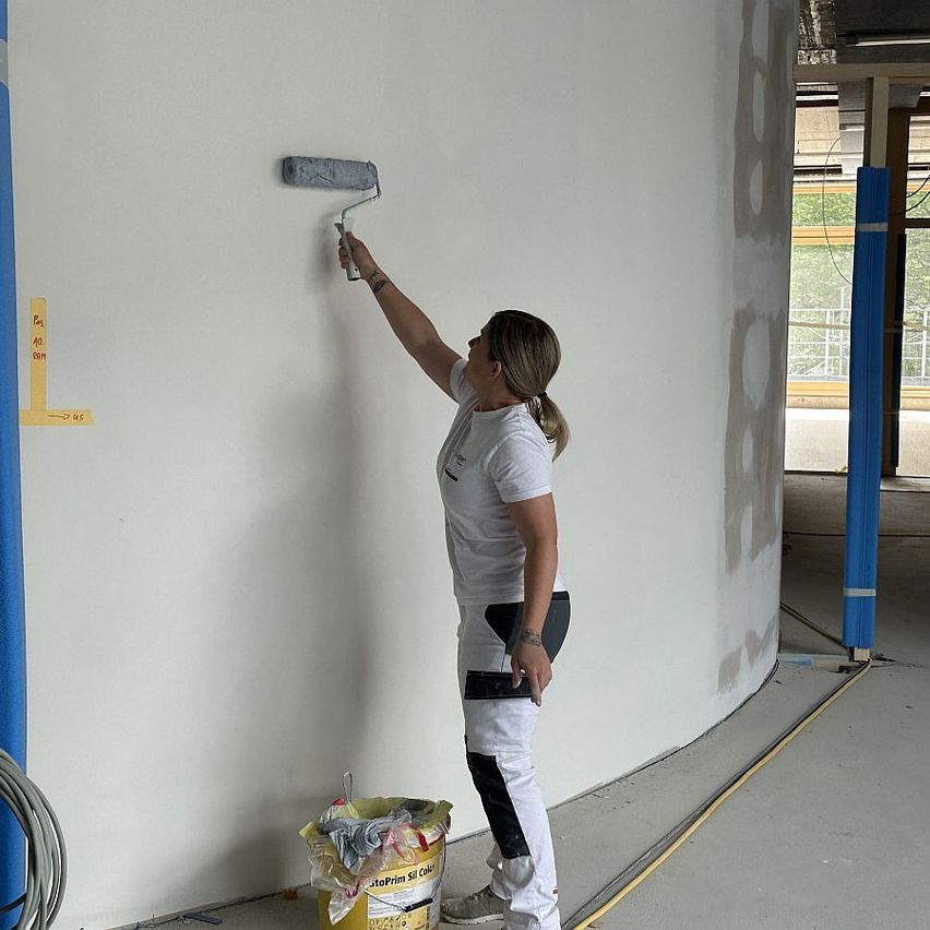 Eine Frau steht an einer Wand und trägt graue Farbe mit einem Farbroller auf. Sie trägt ein weißes Shirt, Hose und Turnschuhe. In der Nähe sind ein gelber Eimer und ein gelbes Klebeband.