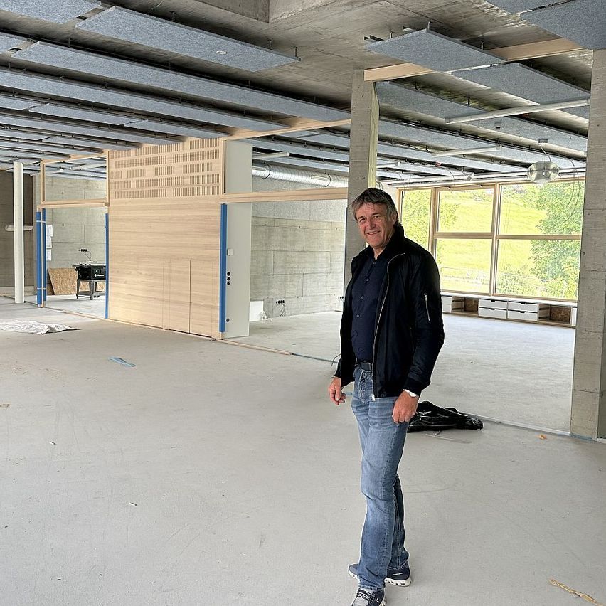 Ein Mann steht in einem unfertigen Raum mit Betonwänden, Holzpaneelen und großen Fenstern. Er lächelt für die Kamera.