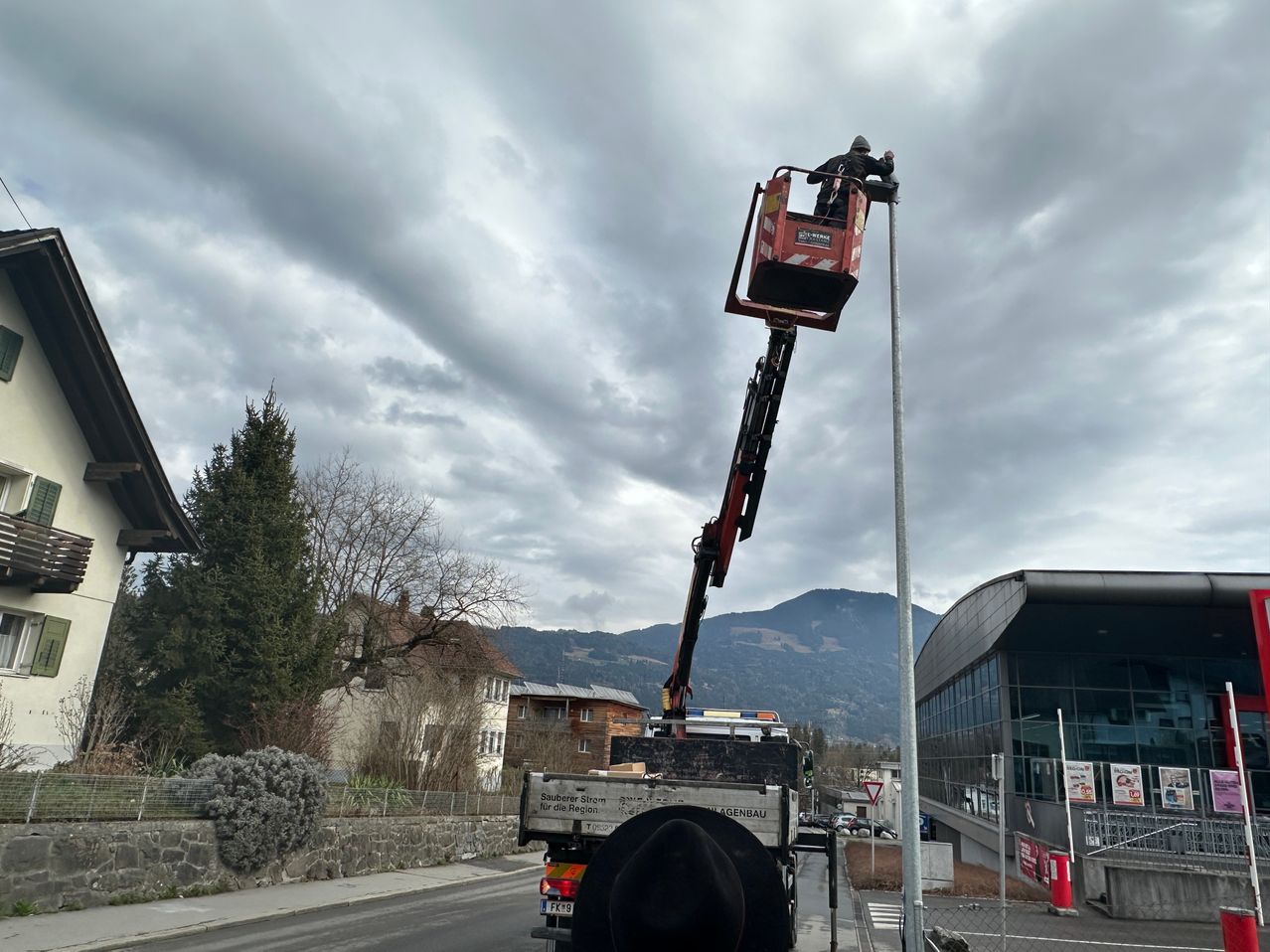 Ein Mann in einer Hebebühne am Straßenrand mit einem bewölkten Himmel und Bergen im Hintergrund.