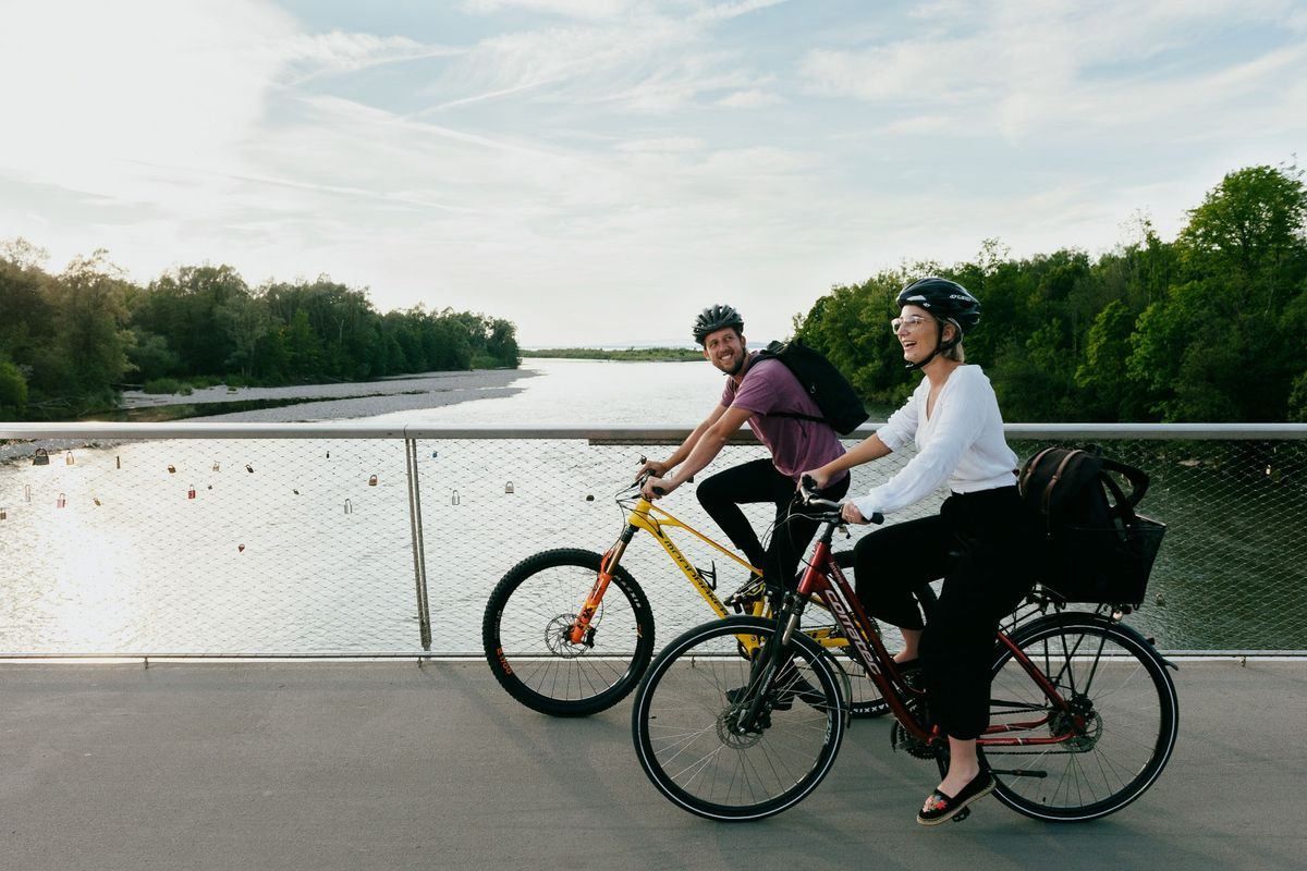 Zwei Personen fahren auf einem Betonweg am Fluss mit Bäumen und einer bewölkten Himmelsscheibe im Hintergrund mit ihren Fahrrädern.
