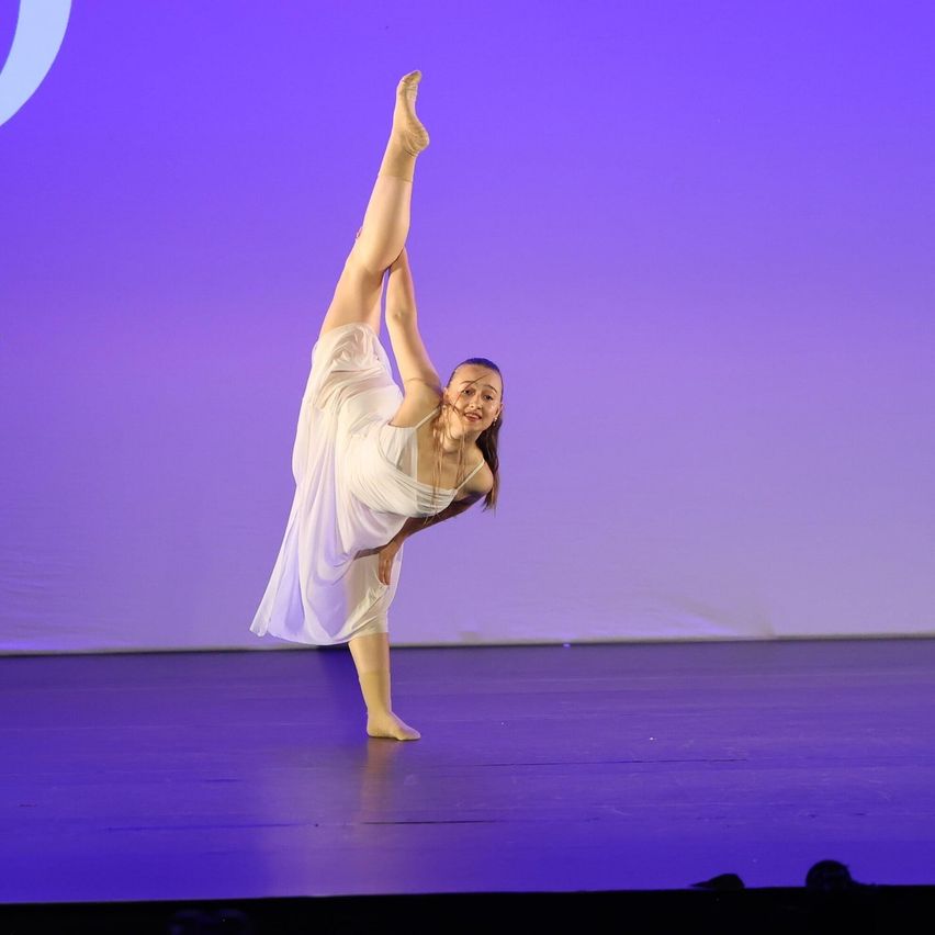 Eine Frau führt eine Ballettpose auf der Bühne aus, trägt ein weißes Kleid und lächelt das Publikum an. Die Bühne ist mit lila Beleuchtung beleuchtet.