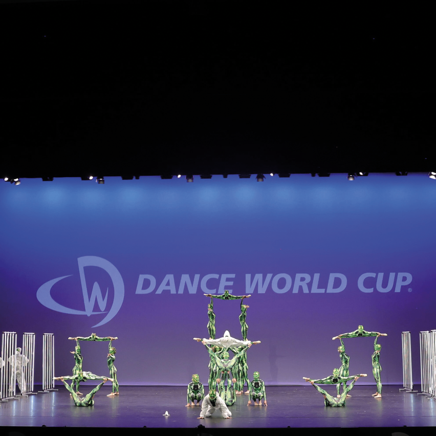 Eine Gruppe von Künstlern in grünen Kostümen führt Akrobatik auf der Bühne beim Dance World Cup auf. Die Bühne ist blau mit dem Logo der Veranstaltung im Hintergrund.