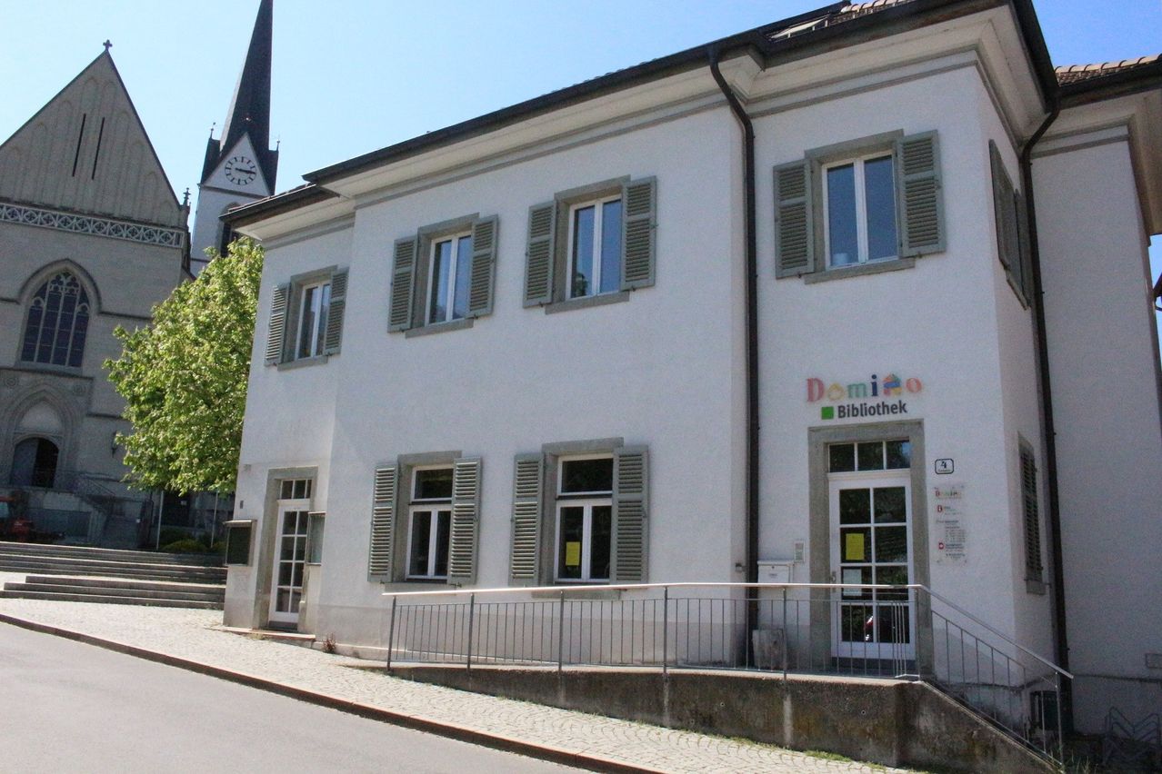 Ein weißes Gebäude mit mehreren Fenstern und Jalousien, einem Glockenturm im Hintergrund und einem Schild, das 'Domizil Bibliothek' besagt.