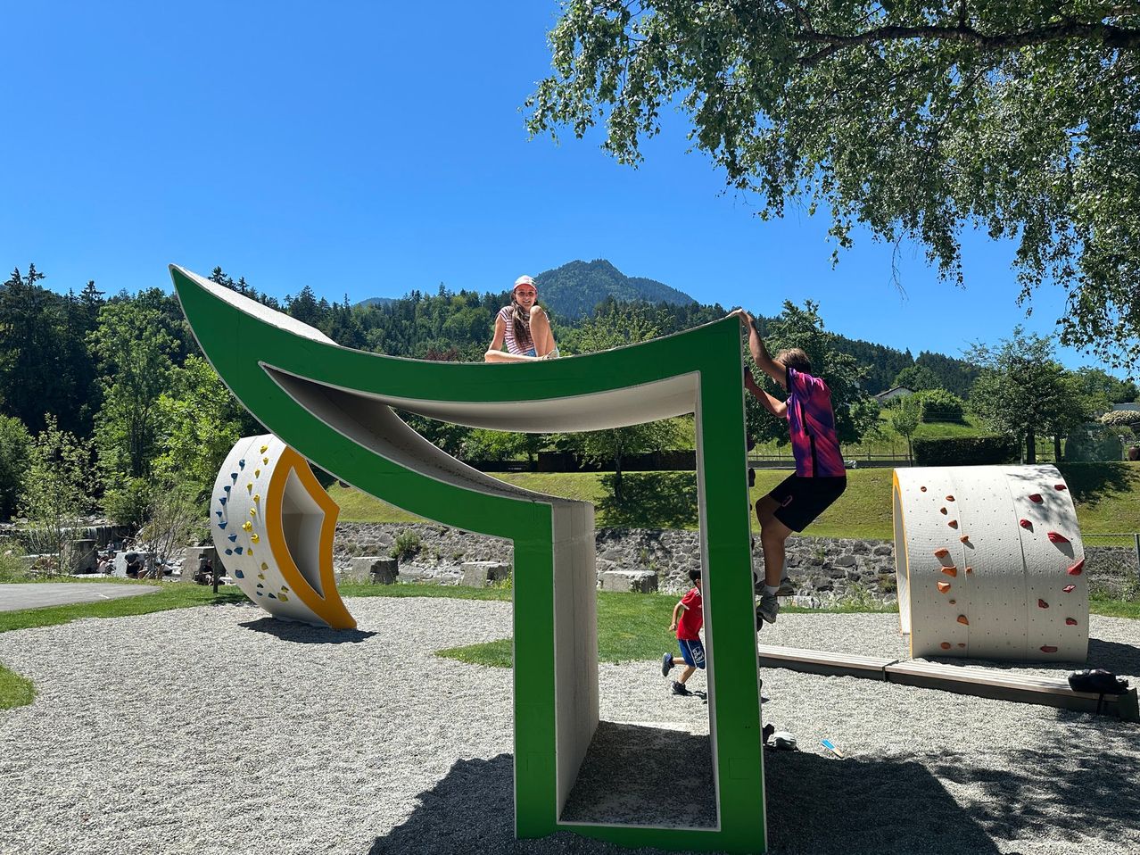 Three people are playing on a large green and white structure in a park with mountains in the background.