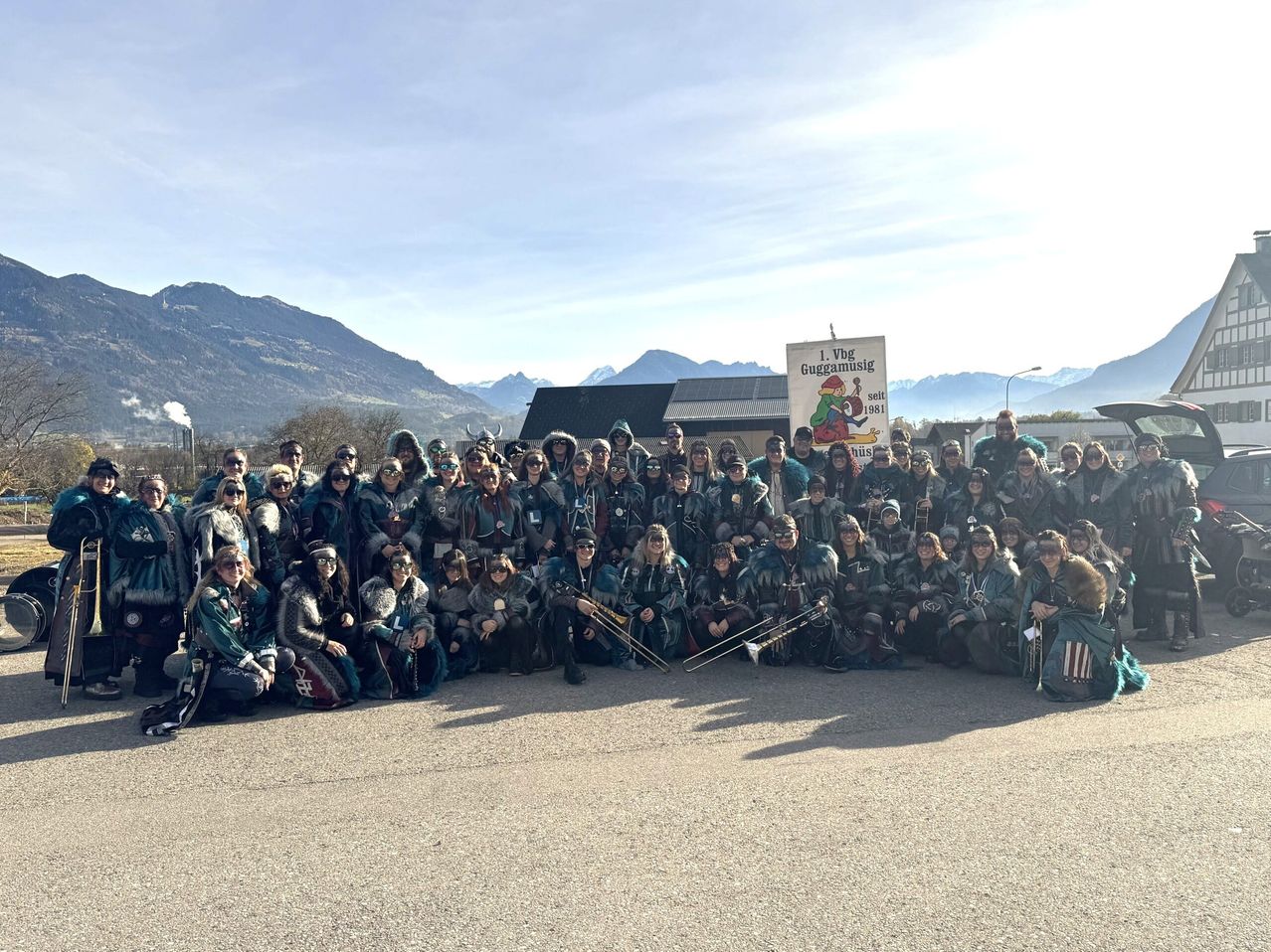 A large group of people dressed in green costumes and masks pose for a photo in front of a mountain range. A banner with a cartoon character and text is behind them.