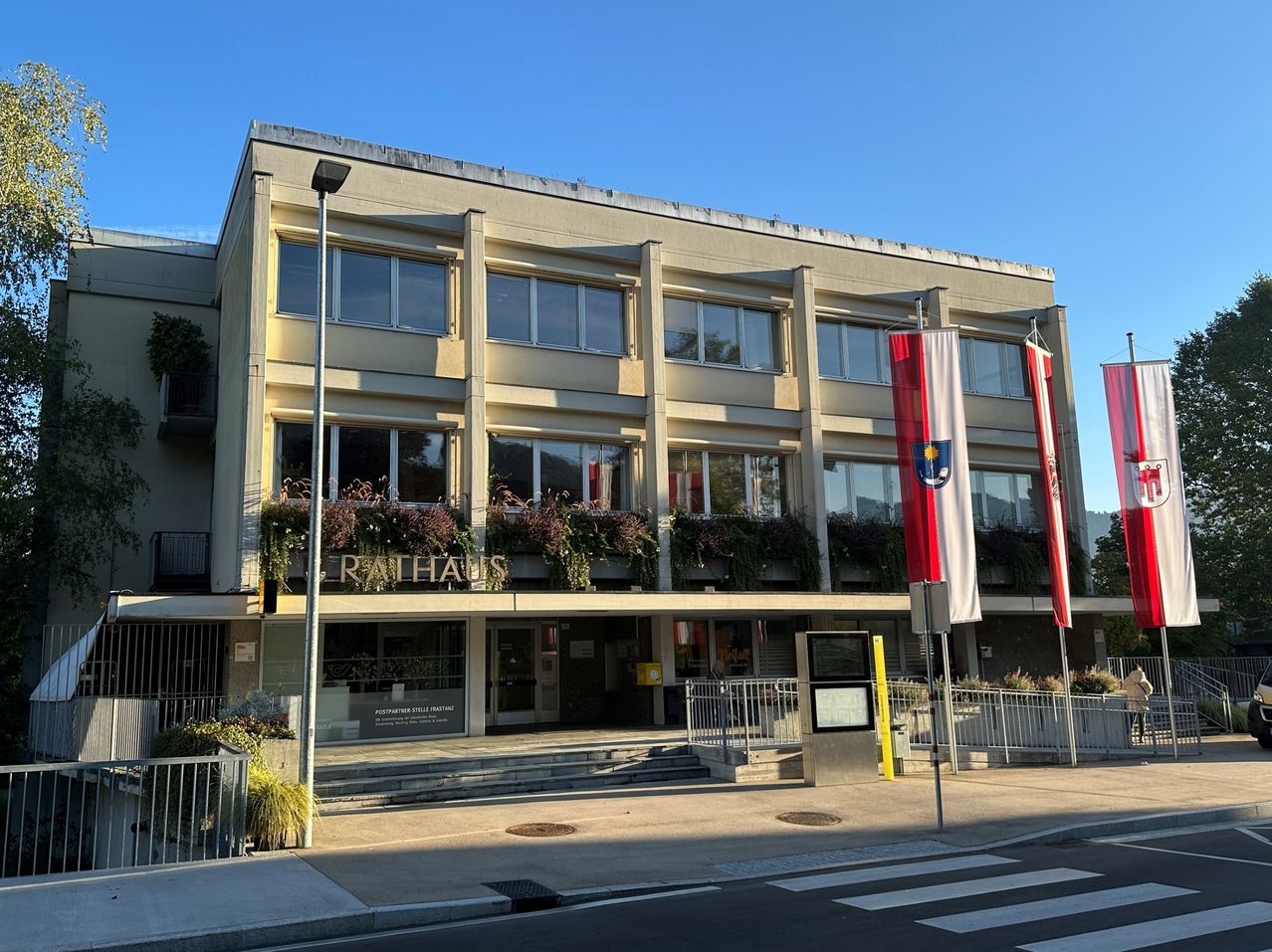 Das Rathausgebäude, ein modernes Bauwerk mit Glasfenstern und einem Balkon, der mit Pflanzen geschmückt ist, steht prominent mit Fahnen davor.