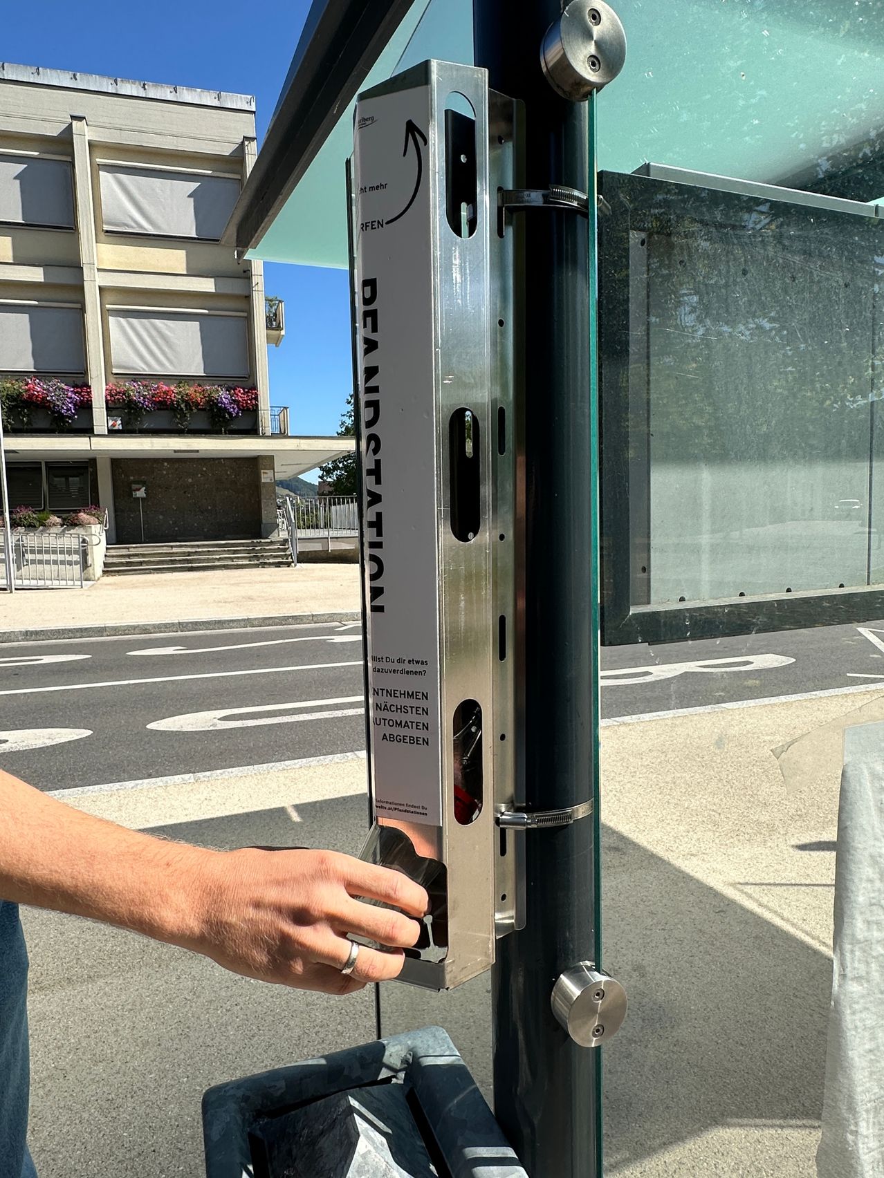 A person is interacting with a metal box attached to a glass door, labeled 'BEARSTATION'. The box contains a lock and a sign that reads 'NTNEHMEN NACHSTEMEN AUTOMATEN ABGEBEN'. The background shows a building with flowers and a road.