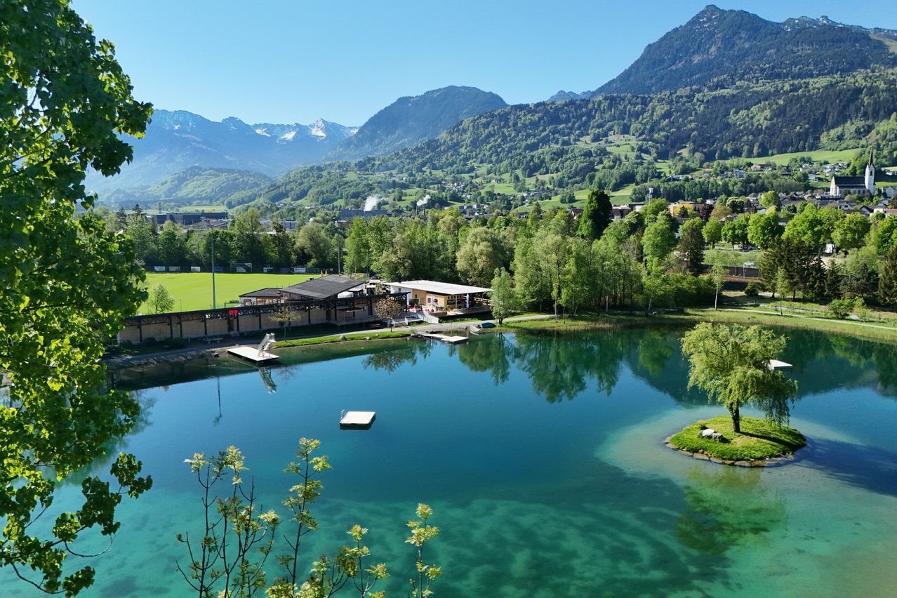 Ein ruhiger See mit Sprungbrett und kleiner Anlegestelle, umgeben von üppigen grünen Bäumen und Bergen im Hintergrund.