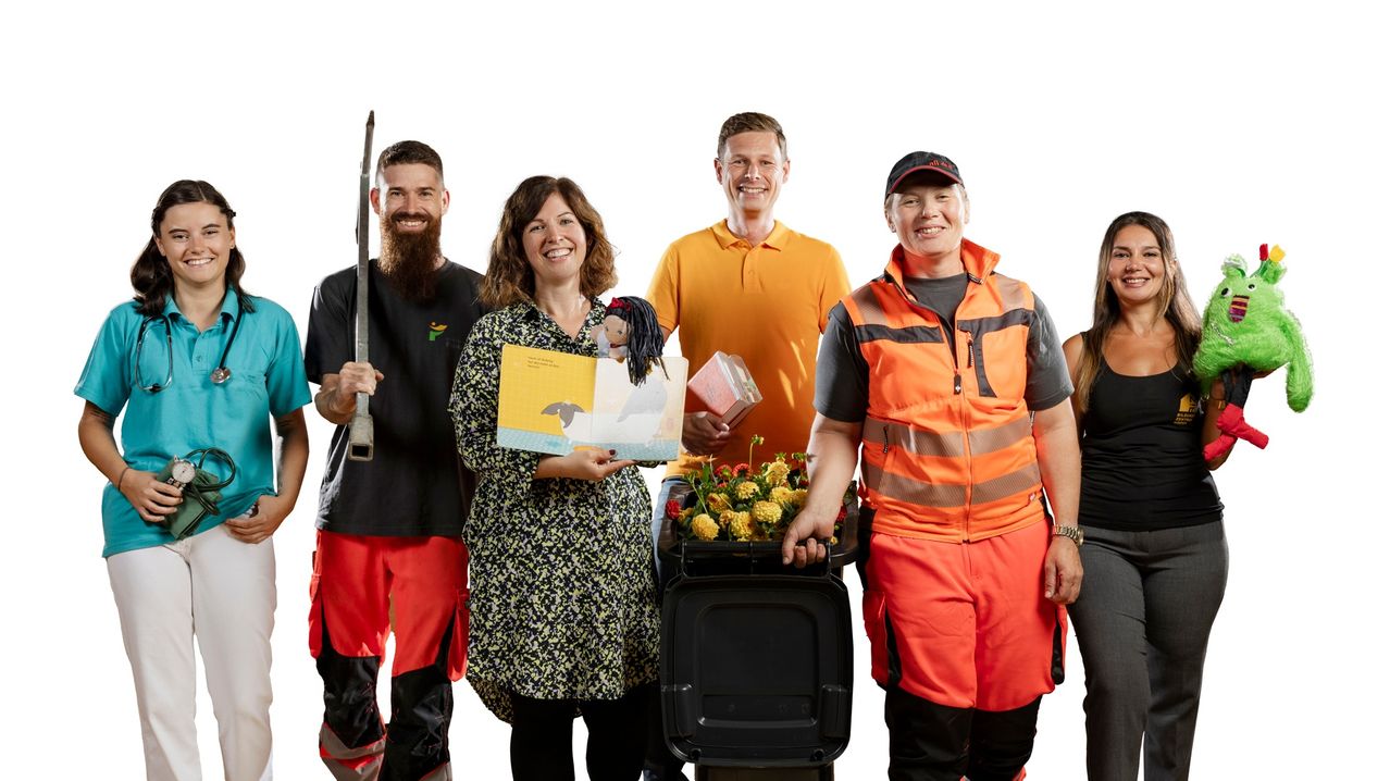 A group of five people standing together, smiling for a photo. One holds a book and doll, another a trash bin, and others hold various items. They wear different colored uniforms.