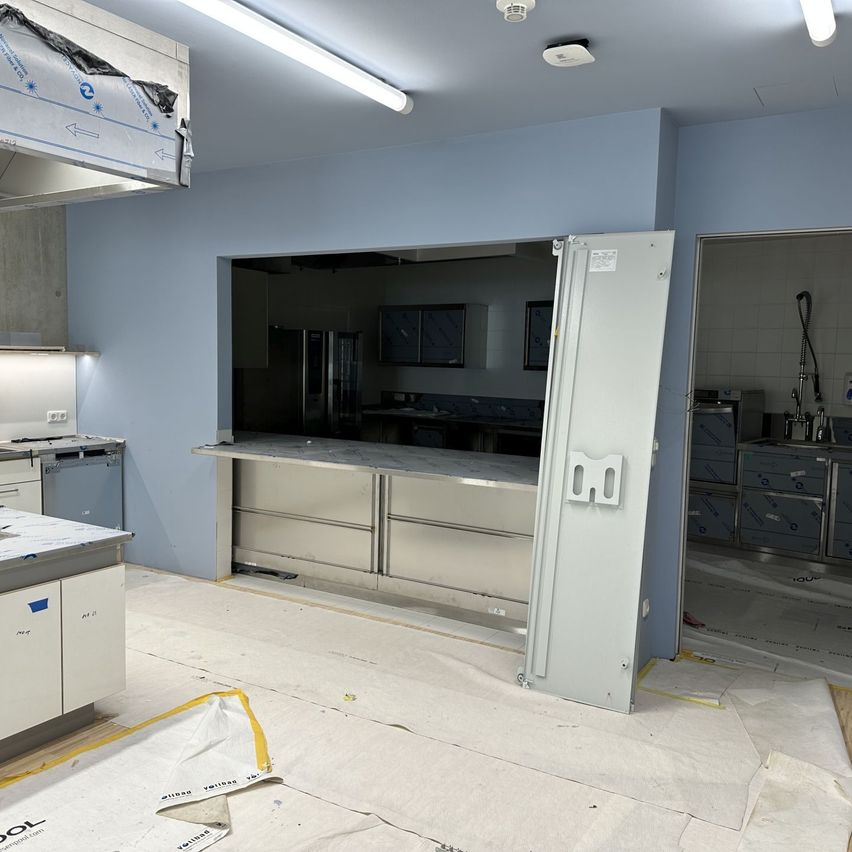 An under-construction kitchen with blue walls, stainless steel cabinets, and an open door. The room is illuminated by ceiling lights.