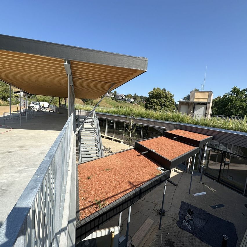 An aerial view of a modern building with a green roof, featuring a metal railing, a staircase, and a car parked in the distance.