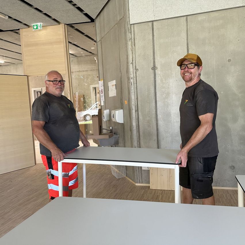 Two men in a room with wooden floors, standing next to a white table, one wearing a hat and glasses, the other in reflective clothing. Behind them, a gray wall with a sink and a car visible through a glass door.