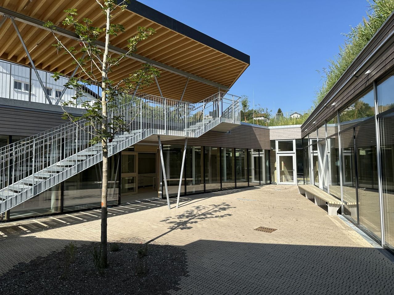 A modern building with a wooden roof and glass walls, featuring a staircase leading to a balcony. A small tree stands in the foreground.