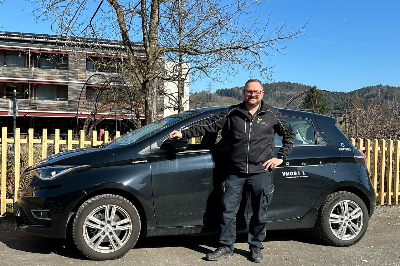 Ein Mann steht neben einem schwarzen Auto mit einem VMOBI L Logo, trägt eine schwarze Jacke und eine Brille, vor einem Gebäude mit einem Baum und einem klaren Himmel.