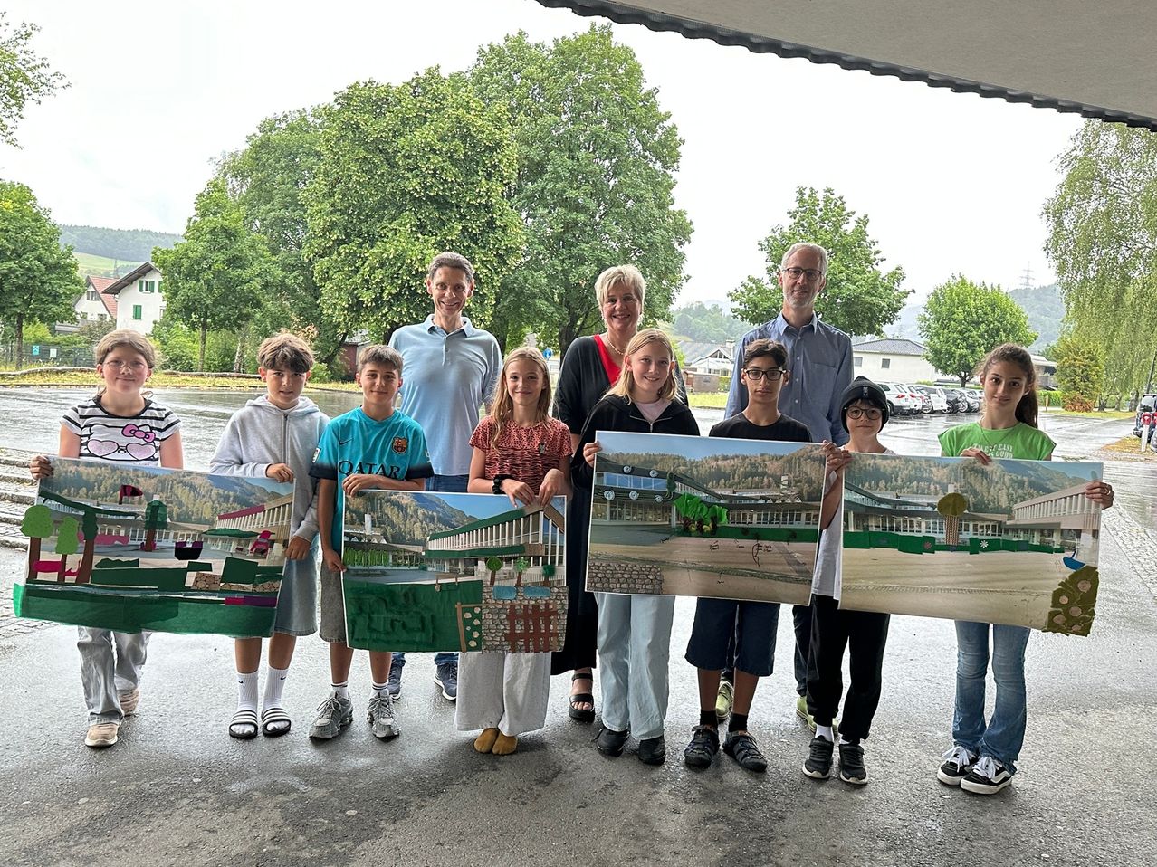 Eine Gruppe von Schülern und Lehrern posiert für ein Foto und hält große Gemälde von Gebäuden, mit Bäumen und einem bewölkten Himmel im Hintergrund.