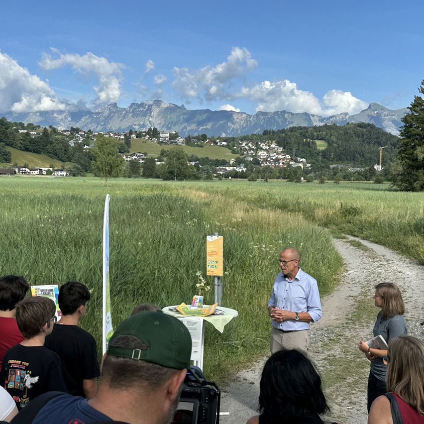 Ein Mann steht auf einem Lehmweg und spricht zu einer Gruppe von Menschen. Hinter ihm steht ein Tisch mit Blumen und einem Schild. Im Hintergrund ein grasiger Feld und Berge.