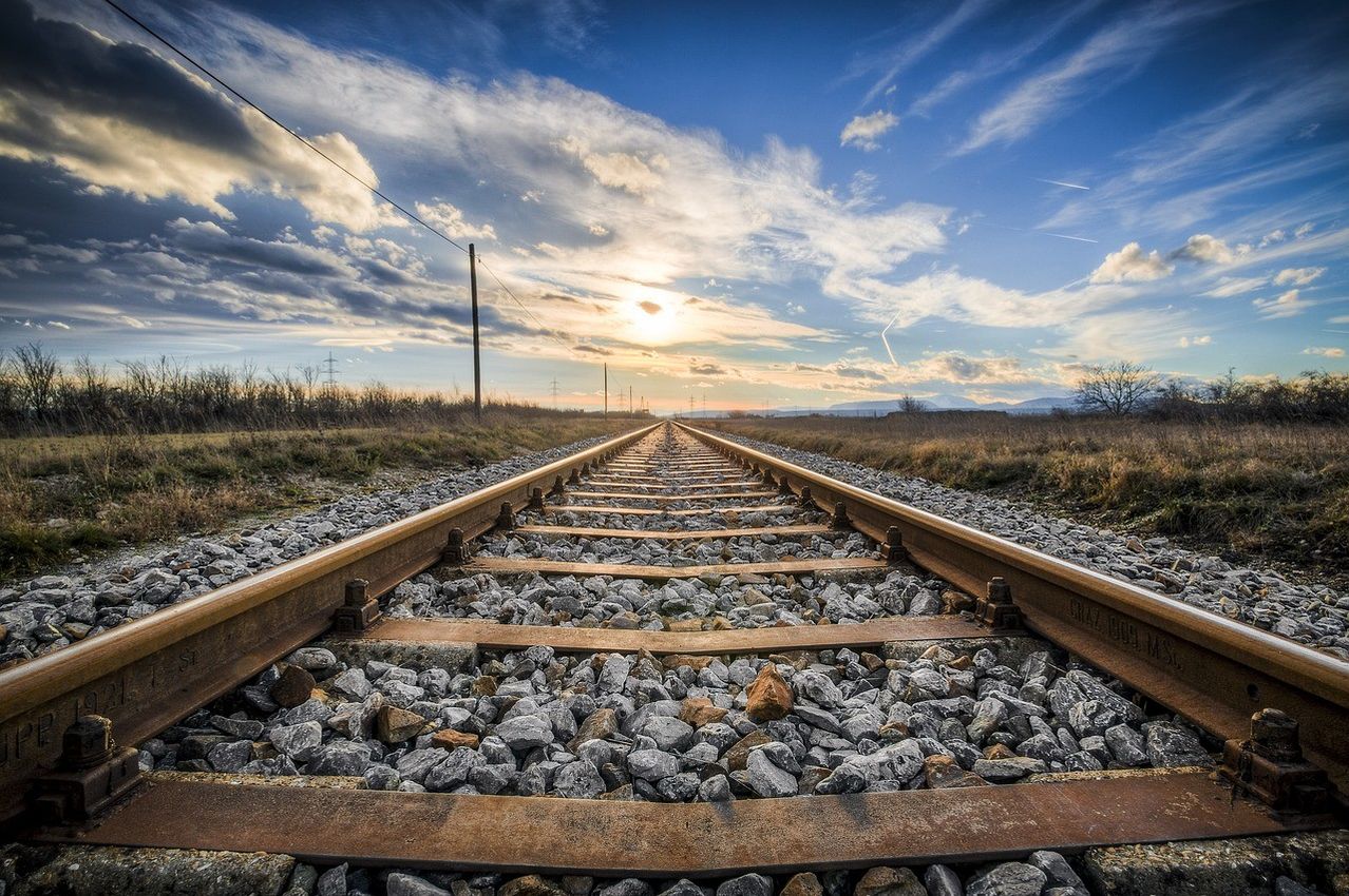 Eine lange Eisenbahnstrecke erstreckt sich unter einem blauen Himmel mit verstreuten Wolken in die Ferne. Die untergehende Sonne wirft ein warmes Licht auf die Kies- und Stahlschienen.