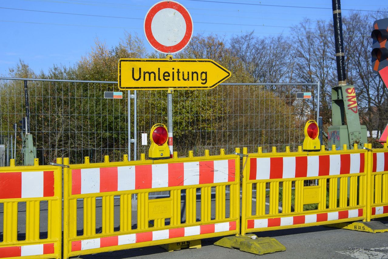 Ein gelbes Straßenschild mit dem Wort 'Umleitung', das eine Umleitung anzeigt. Es ist an einem Metallpfosten befestigt, über dem sich ein rotes und weißes kreisförmiges Schild befindet. Auf dem Pfosten befinden sich zwei rote Lichter. Dahinter steht ein Metallzaun mit Bäumen im Hintergrund.