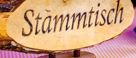 Ein Holzschild mit dem Wort Stammtisch wird auf einem gestreiften Tuch gezeigt. Das Schild ist aus einem Baumstamm geschnitten.