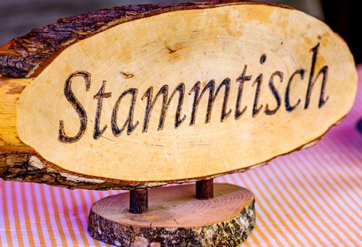 Ein Holzschild mit dem Wort Stammtisch wird auf einem gestreiften Tuch gezeigt. Das Schild ist aus einem Baumstamm geschnitten.