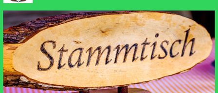 Ein Holzschild mit Stammtisch darauf, stehend auf einem gestreiften Tischtuch. Das Schild ist auf einem grünen Hintergrund.