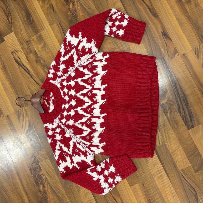 A red knitted sweater with white snowflake patterns is hung on a wooden floor. The sweater is folded and placed on a hanger.