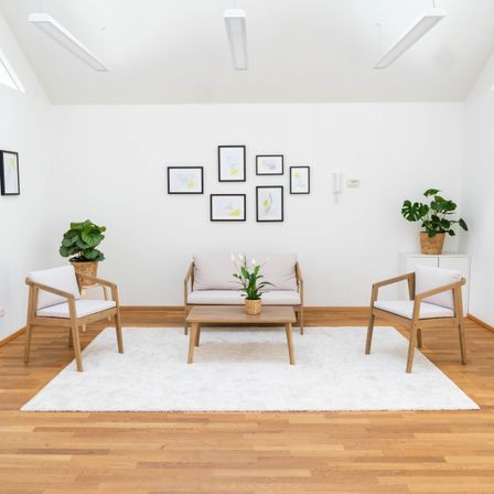 Ein modernes Wohnzimmer mit weißen Wänden und Holzböden. Eine weiße Couch mit einem Blumentopf steht auf einem Teppich in der Mitte. Zwei Stühle sind auf beiden Seiten. Gerahmte Bilder schmücken die Wand.