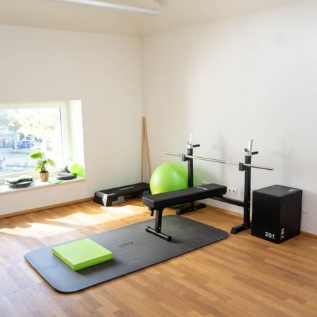 Ein Heim-Fitnessstudio mit einer Bankpresse, einem Gewichtsständer und einem Schaumstoffblock auf einer Yogamatte. Ein grüner Ball und eine Topfpflanze sind auf der Fensterbank.