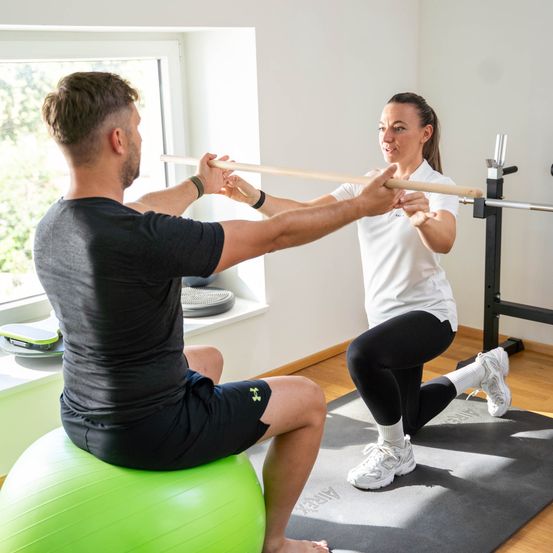 Ein Mann und eine Frau trainieren gemeinsam im Fitnessstudio. Der Mann sitzt auf einem grünen Übungsball, während die Frau ihm beim Dehnen mit einer Holzstange hilft.