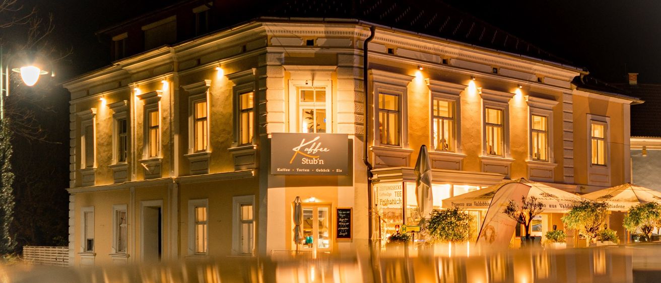 Ein Gebäude mit mehreren Fenstern ist nachts hell beleuchtet, mit Reflexionen auf dem Boden darunter.