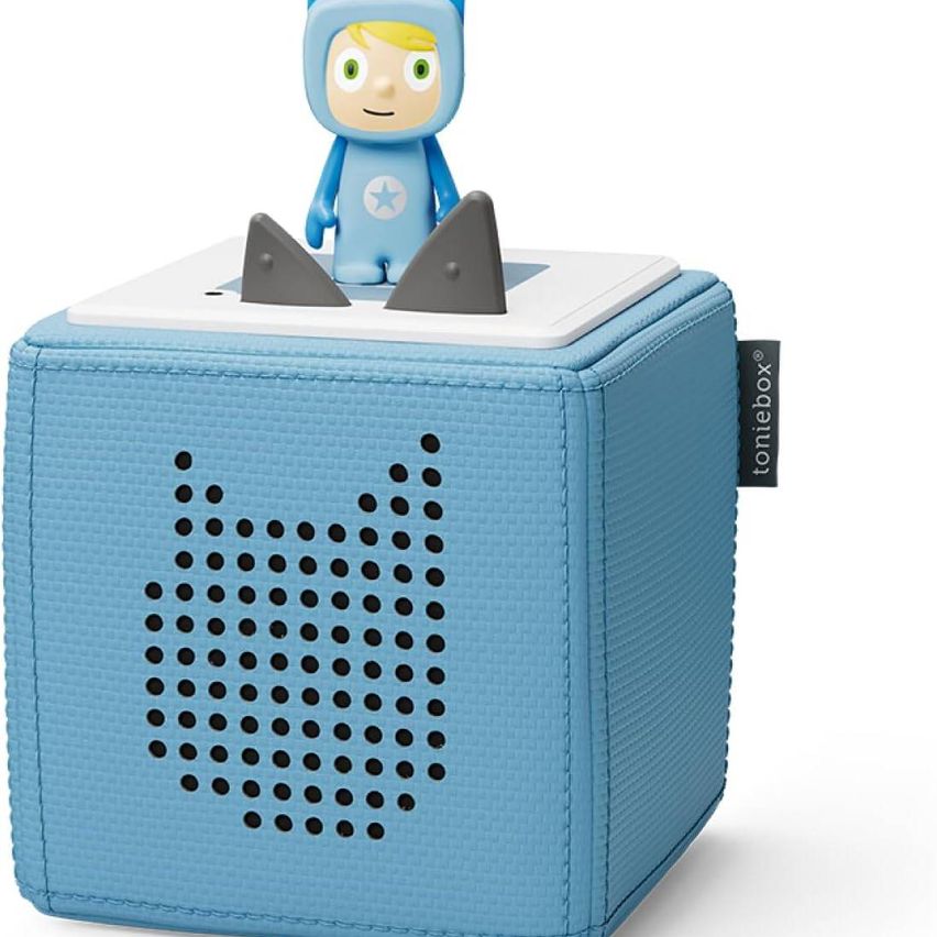 Ein hellblauer Würfel-Lautsprecher mit strukturierter Oberfläche und einer kleinen Spielzeugfigur oben drauf. Das Spielzeug ist blau mit einem Stern auf der Brust.