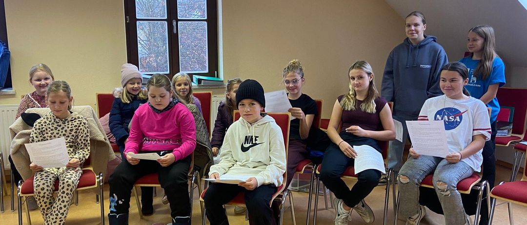 Eine Gruppe von Kindern in einem Klassenzimmer sitzt auf Stühlen, hält Papiere in den Händen und scheint an einer Lernaktivität teilzunehmen. Einige tragen Winterkleidung.