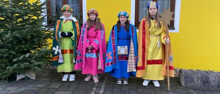 Vier Mädchen in farbenfrohen Kostümen posieren vor einem gelben Gebäude mit Fenstern und einem Weihnachtsbaum. Jedes von ihnen hält eine sternförmige Requisite in der Hand.