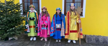 Vier Frauen in farbenfrohen Kostümen stehen vor einem gelben Gebäude mit Fenstern. Sie posieren für ein Foto.