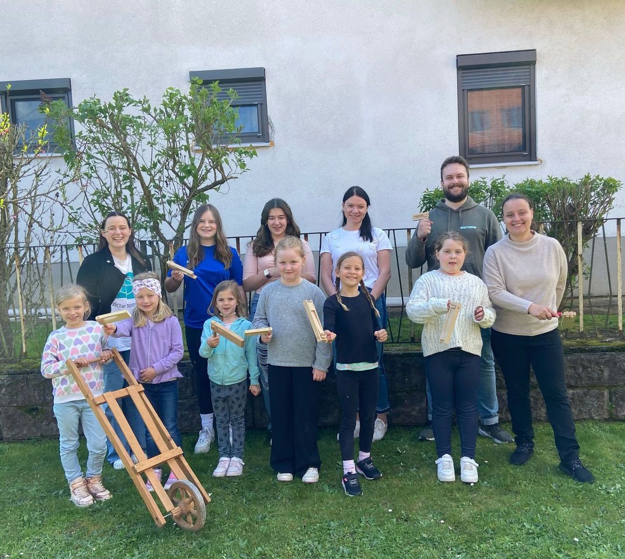 Eine Gruppe von Erwachsenen und Kindern steht draußen und posiert für ein Foto. Sie halten Holzobjekte und einige lächeln. Im Hintergrund ist ein Gebäude mit Pflanzen zu sehen.