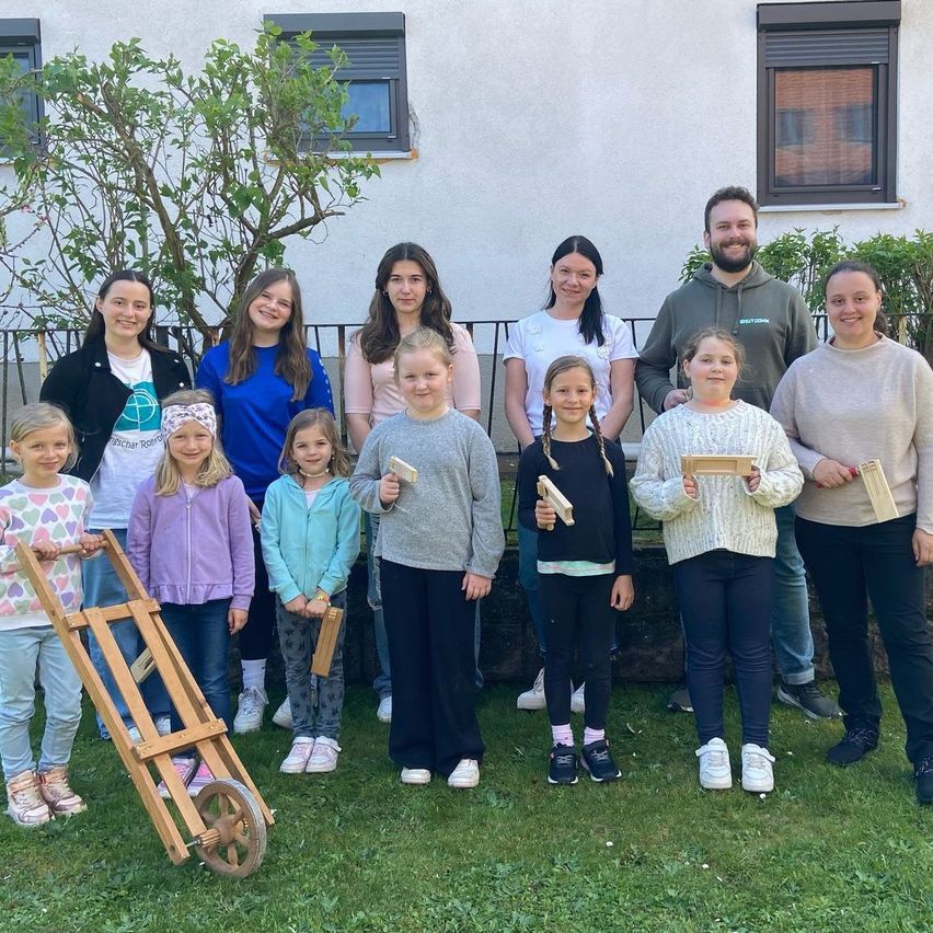 Eine Gruppe von Kindern und zwei Erwachsenen posiert für ein Foto vor einem Gebäude. Sie halten alle Holzwerkzeuge in den Händen.
