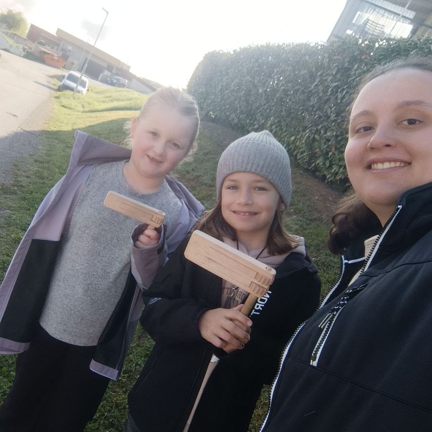 Drei Mädchen stehen vor einer Hecke, lächeln und halten Holzpaddel in den Händen, die wahrscheinlich für ein Spiel verwendet werden. Ein Mädchen trägt eine graue Mütze, ein anderes eine Jacke mit lila und schwarzen Akzenten und das dritte eine schwarze Jacke.