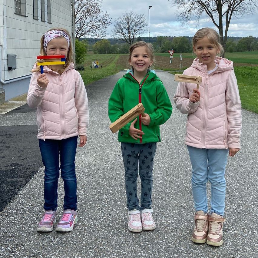Drei junge Mädchen stehen auf einem gepflasterten Weg und halten Holzspielzeug in den Händen. Das Mädchen auf der linken Seite trägt eine rosa Jacke und blaue Jeans. Das Mädchen in der Mitte trägt eine grüne Jacke mit Reißverschluss. Das Mädchen auf der rechten Seite hat eine rosa Jacke mit Kapuze.