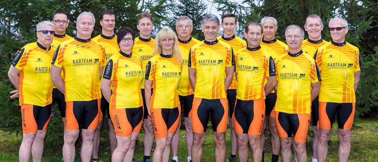 Eine Gruppe von Menschen, die in übereinstimmenden gelben und schwarzen Radsporttrikots gekleidet sind, steht in einer Reihe auf einem Rasenfeld, mit Bäumen im Hintergrund.