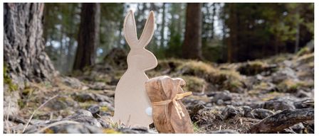 Eine hölzerne Hasenfigur steht auf dem Boden in einem Waldgebiet, mit einem weißen Ei und einem hölzernen Geschenkbeutel in der Nähe.