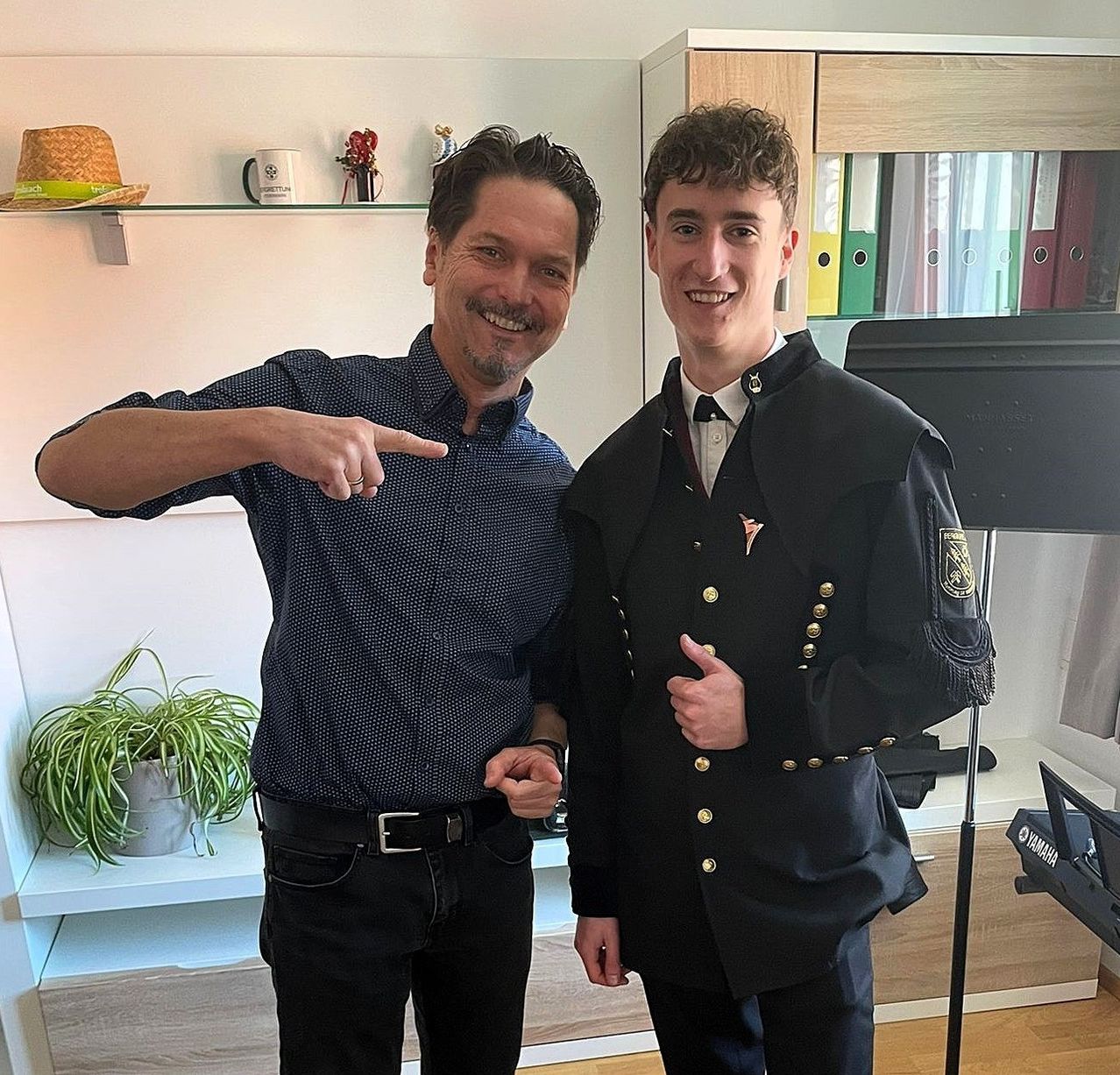 Zwei Männer posieren für ein Foto; einer zeigt mit dem Finger, während er lächelt, der andere in Uniform. Sie stehen neben einem Regal mit Hut, Tasse und Ordnern.