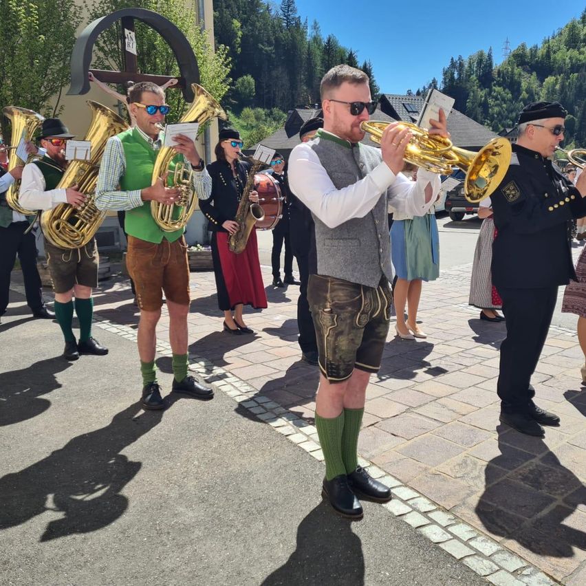 Eine Blaskapelle in traditioneller bayerischer Kleidung spielt Instrumente auf einer Kopfsteinpflasterstraße in den Bergen. Ein Mann spielt Trompete, während ein anderer Tuba spielt.
