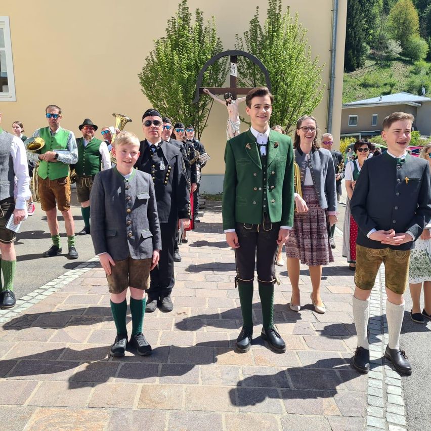 Eine Gruppe von Menschen in traditioneller Kleidung steht draußen, wahrscheinlich bei einer Veranstaltung. Ein Mann in einem grünen Jackett lächelt. Hinter ihm halten andere Musikinstrumente. Ein Kreuz und ein Gebäude sind im Hintergrund zu sehen.