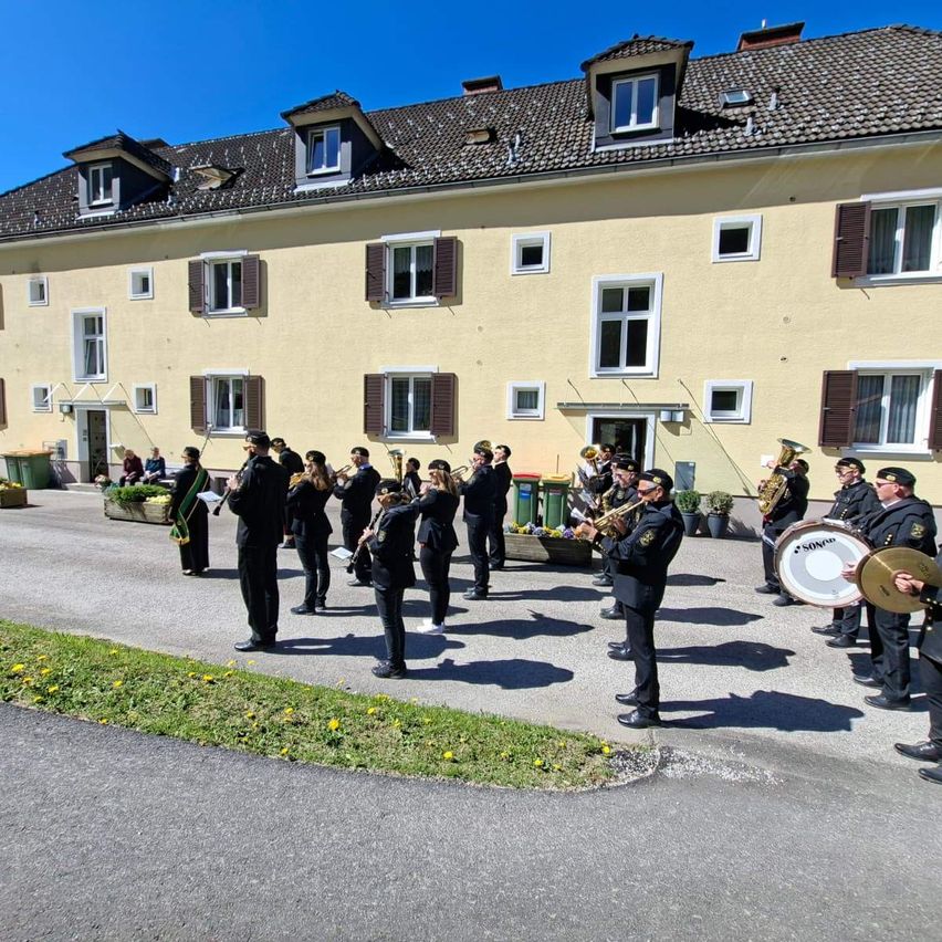 Ein Blasorchester übt vor einem gelben Gebäude mit mehreren Fenstern und Topfpflanzen draußen. Einige Mitglieder spielen Trompeten, während andere Trommeln spielen.