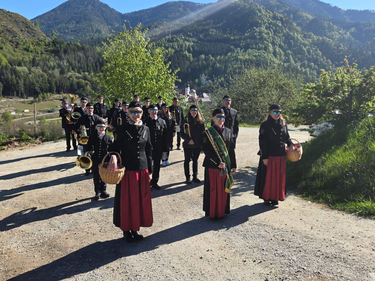 Eine Gruppe von Musikern in traditioneller Kleidung steht auf einem Schotterweg und hält Musikinstrumente in der Hand, mit Bergen im Hintergrund.