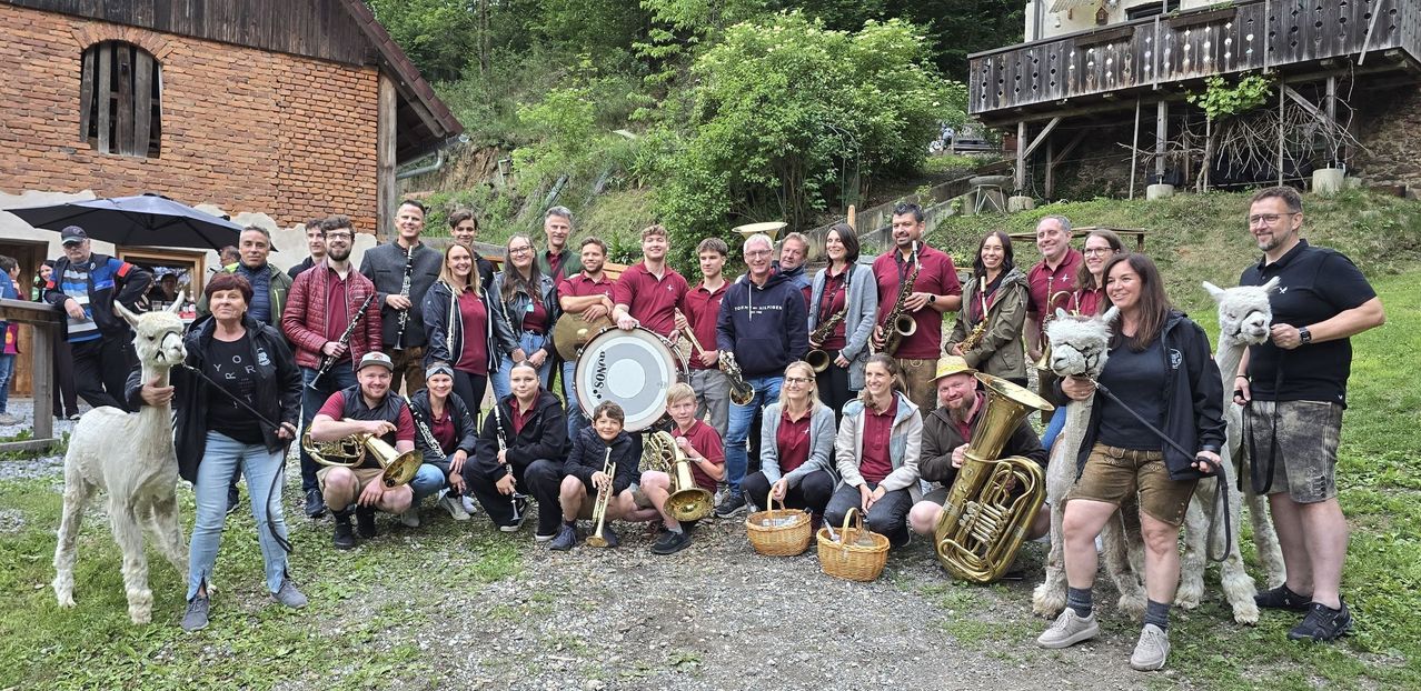 Eine Gruppe von Musikern in roten Hemden posiert mit ihren Instrumenten für ein Foto. Sie sind von Bäumen und einem Holzhaus umgeben.