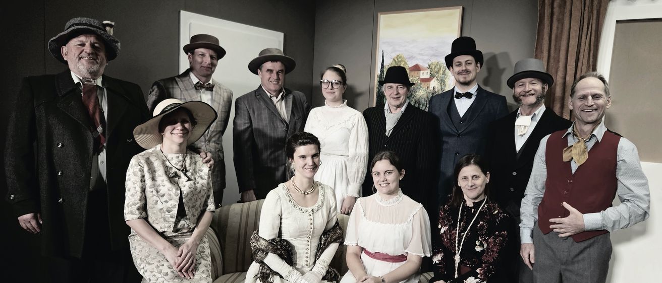 Eine Gruppe von Menschen in Vintage-Kleidung posiert für ein Schwarz-Weiß-Foto. Ein Gemälde eines Hauses ist hinter ihnen.