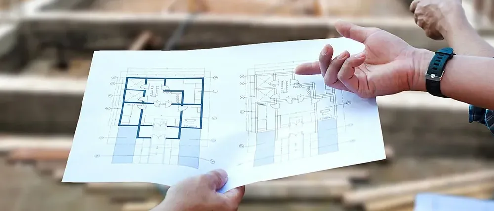 Hände halten ein Papier mit einem Grundriss und zeigen darauf, während ein verschwommener Bauplatz im Hintergrund ist.